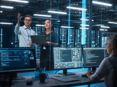 Female e-Business Entrepreneur and Male IT Specialist Discussing Something and Use Laptop in Big Data Center Server Room. Work on Web Services Cloud Computing SAAS Concept